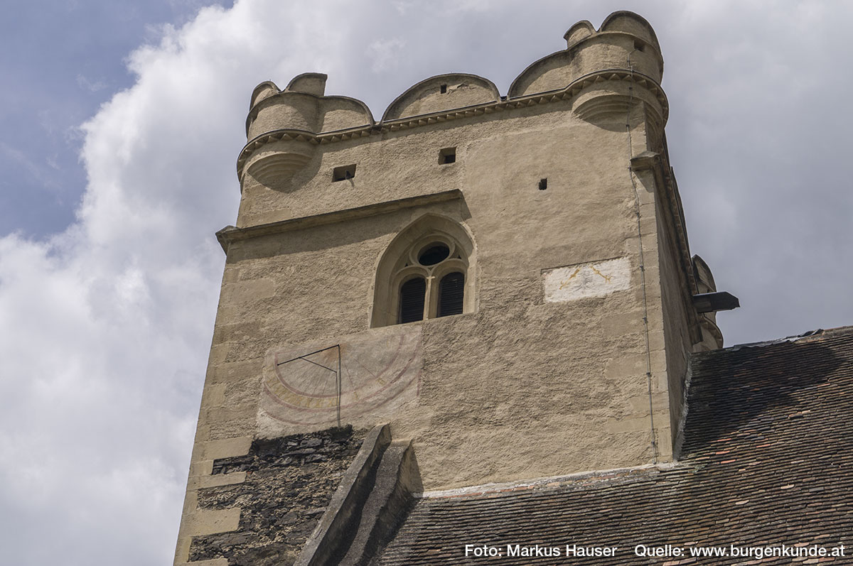 Der Turm der Wehrkirche mit kleinen runden Ecktürmchen, Rundzinnen und einem gotischen Fenster im oberen Bereich. Darunter eine Sonnenuhr und neben den Fenster sind die Füße eines Adlers zu sehen (vll. der Doppeladler?).