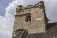 Der Turm der Wehrkirche mit kleinen runden Ecktürmchen, Rundzinnen und einem gotischen Fenster im oberen Bereich. Darunter eine Sonnenuhr und neben den Fenster sind die Füße eines Adlers zu sehen (vll. der Doppeladler?).